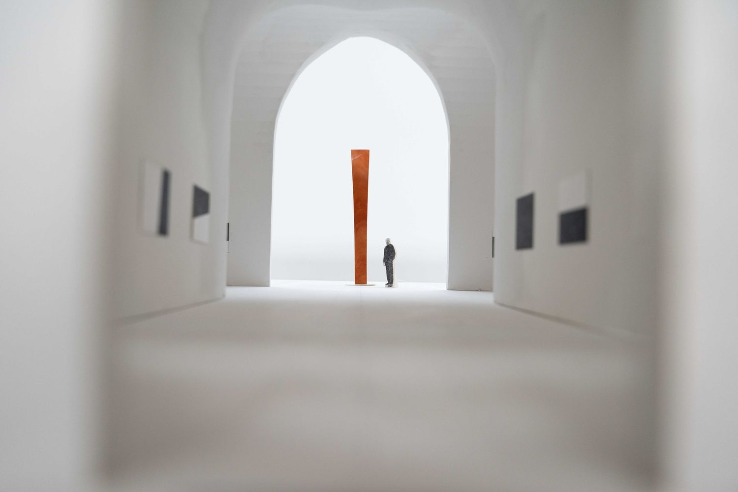 Person standing before tall rust-colored sculpture in white arched gallery corridor.