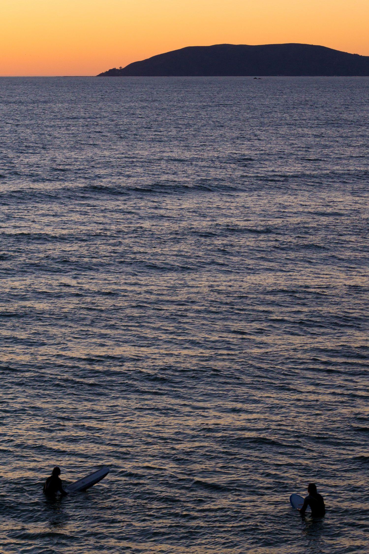 Two surfers silhouetted in calm ocean water at sunset with island visible on horizon.