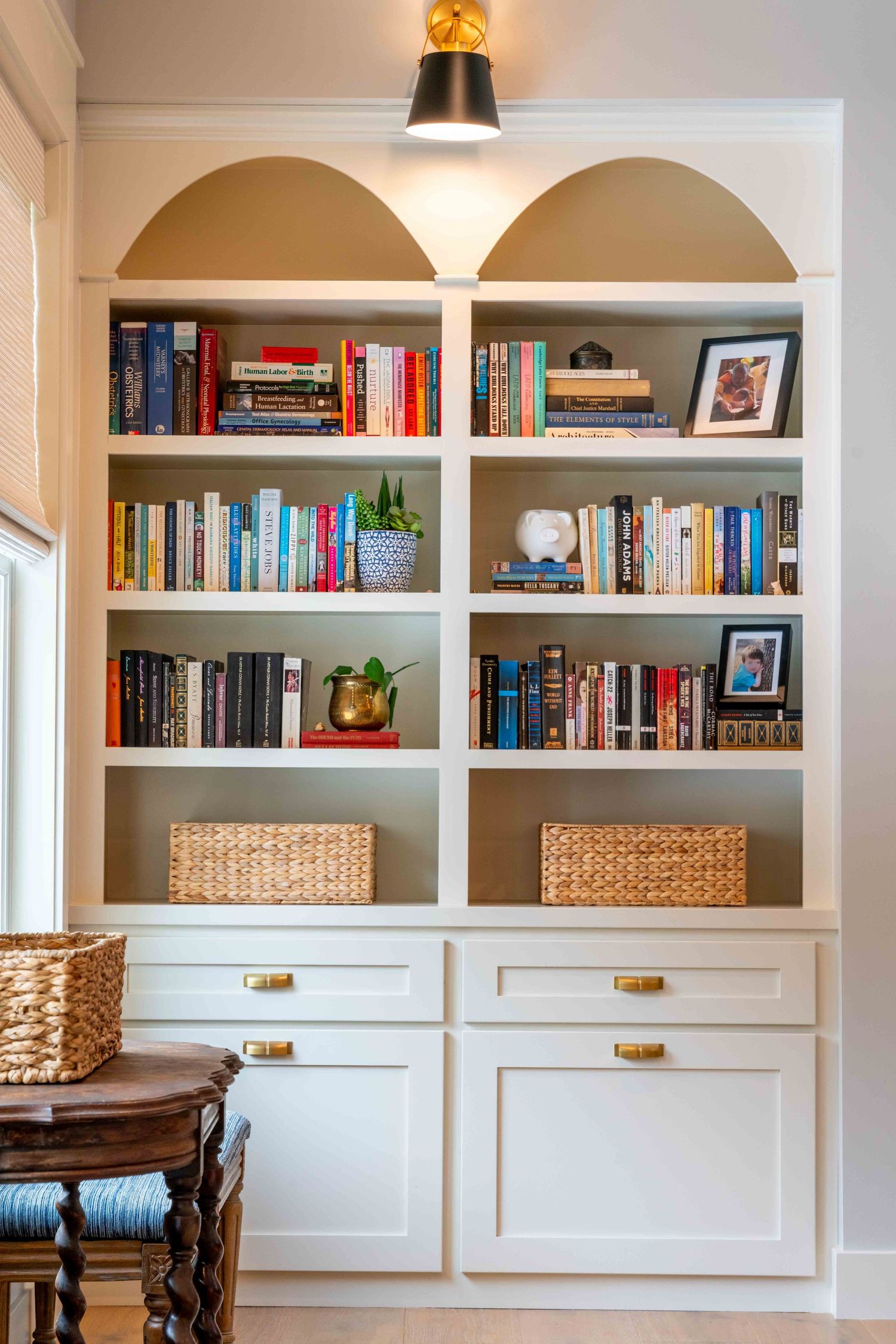 Built-in white bookshelf with arched details, brass hardware, wicker baskets, and colorful books.