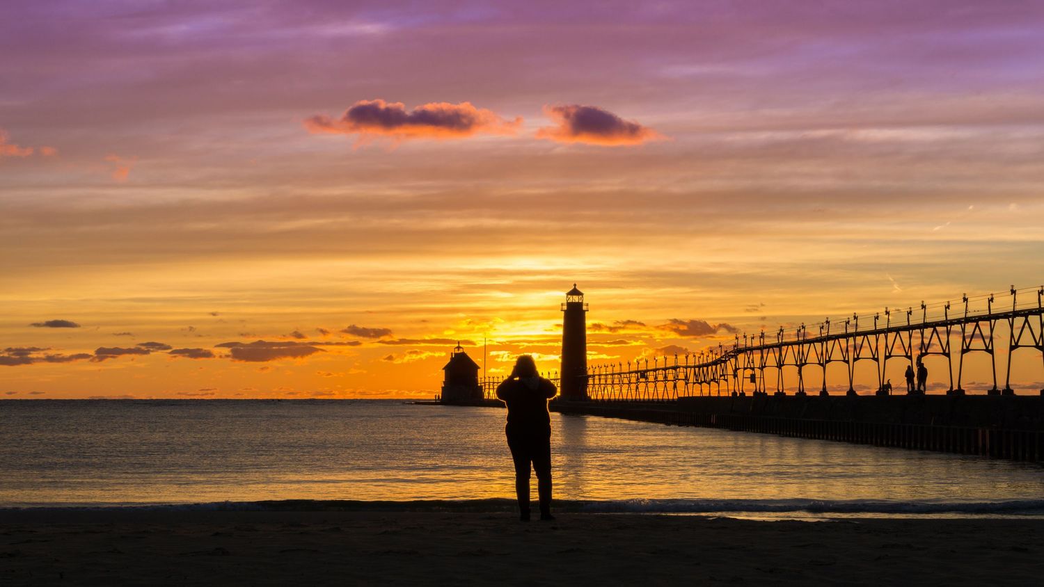 Silhouette of a person standing on a beach at golden sunset beside a lighthouse and illuminated pier.