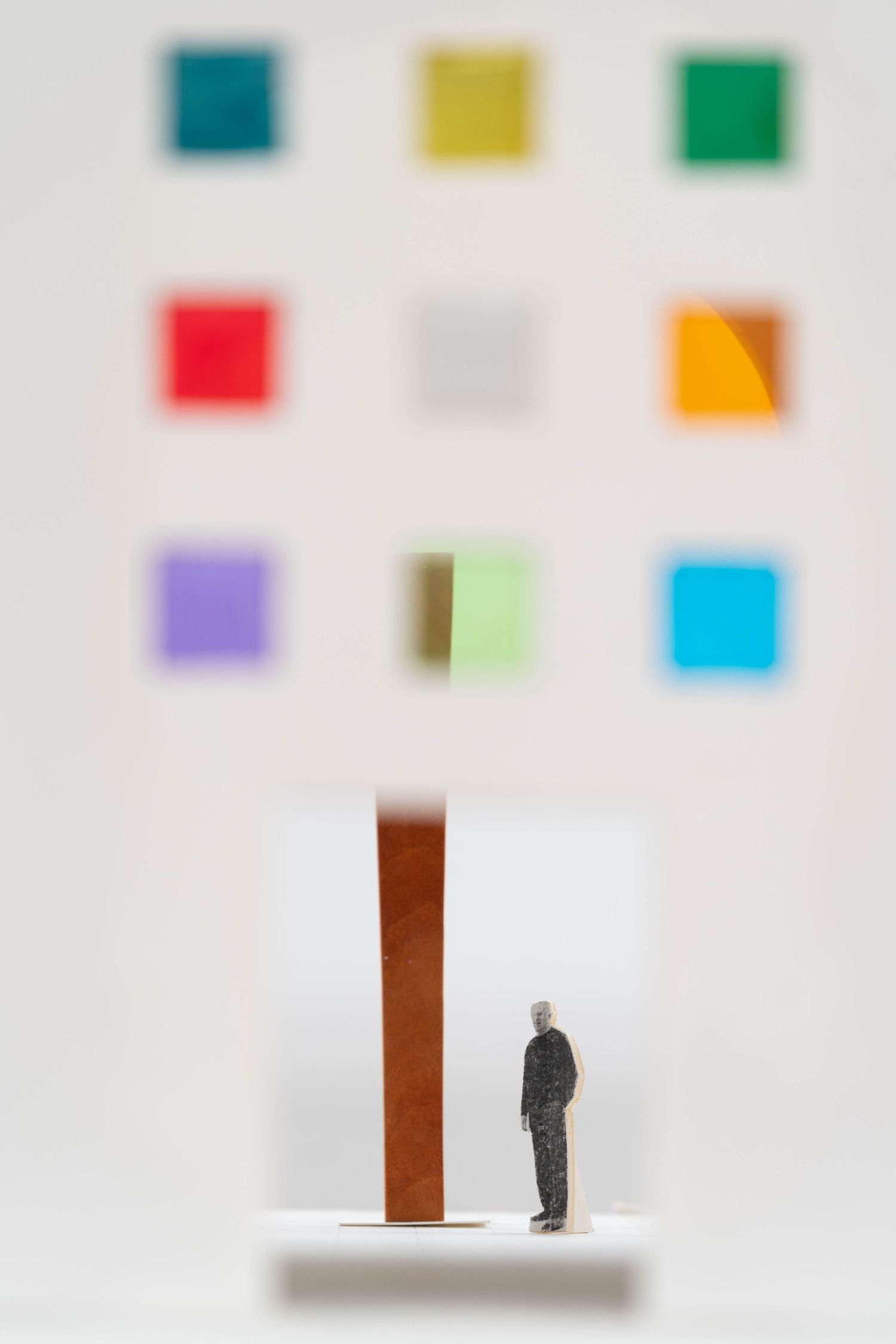 Miniature figure standing beside tall brown pillar with colorful square paintings behind.