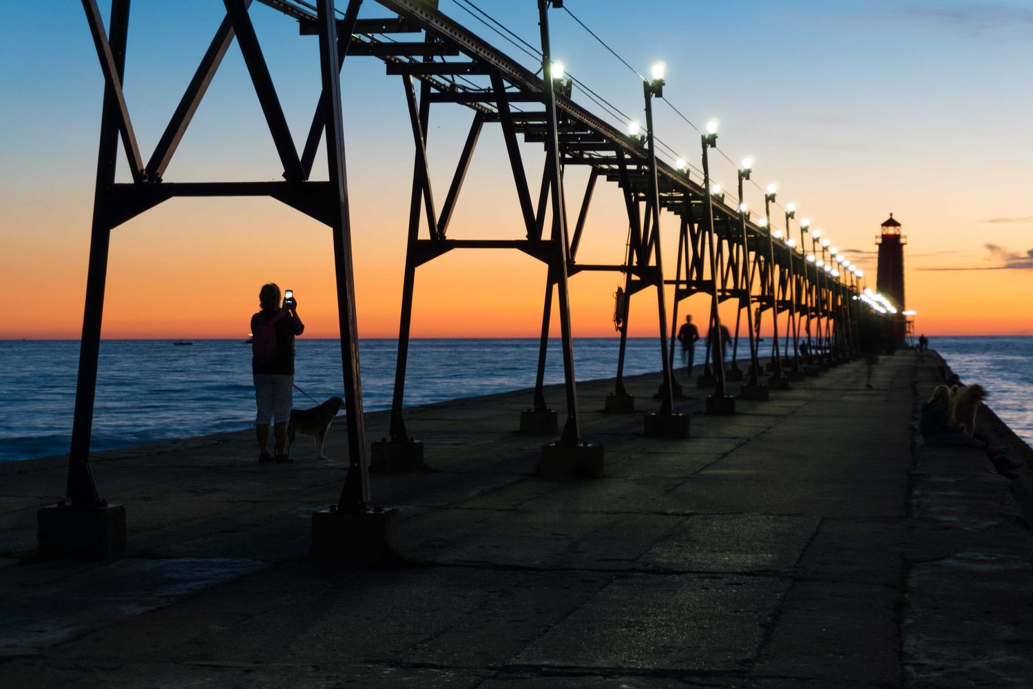 Silhouette of a person standing on a pier with glowing lanterns at sunset, with a lighthouse in the background.