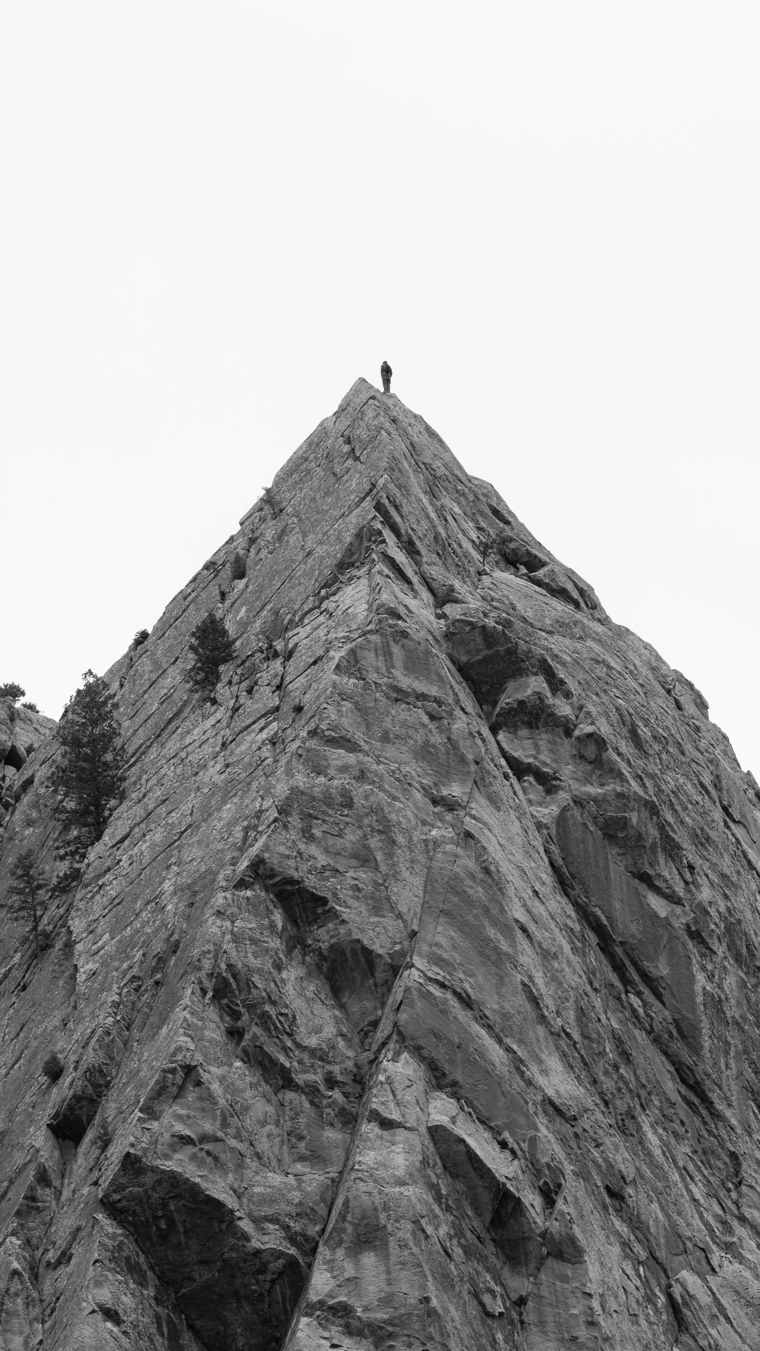 Tiny figure stands atop a dramatic sharp rocky mountain peak against a pale sky.