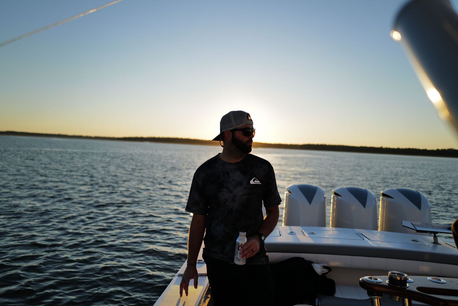 Man in a cap and sunglasses sitting on a boat during a golden sunset over calm water.