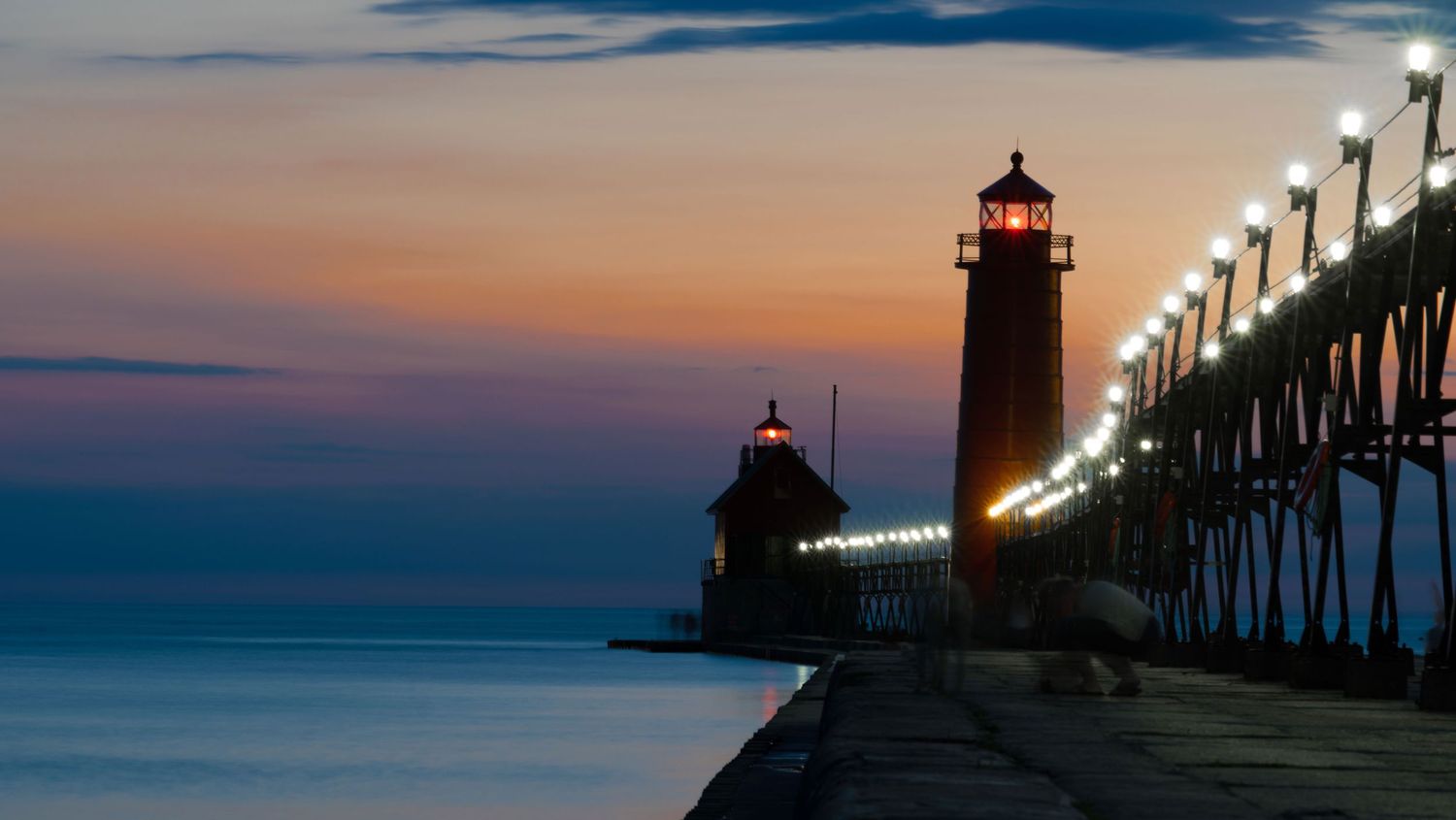 Twin lighthouses with glowing pier lights reflected on calm water at twilight in Grand Haven.