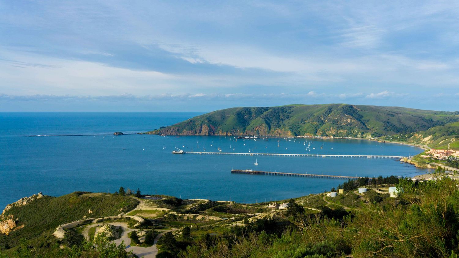 Aerial panoramic view of a blue bay with marina, green hills, and Mediterranean coastline under cloudy sky.