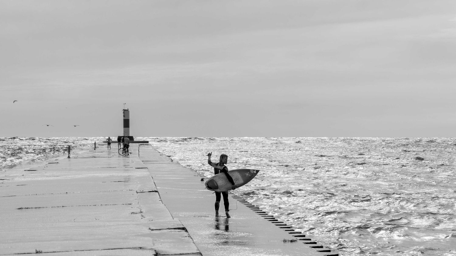 Surfer holding board on a concrete pier with icy waves in black and white.
