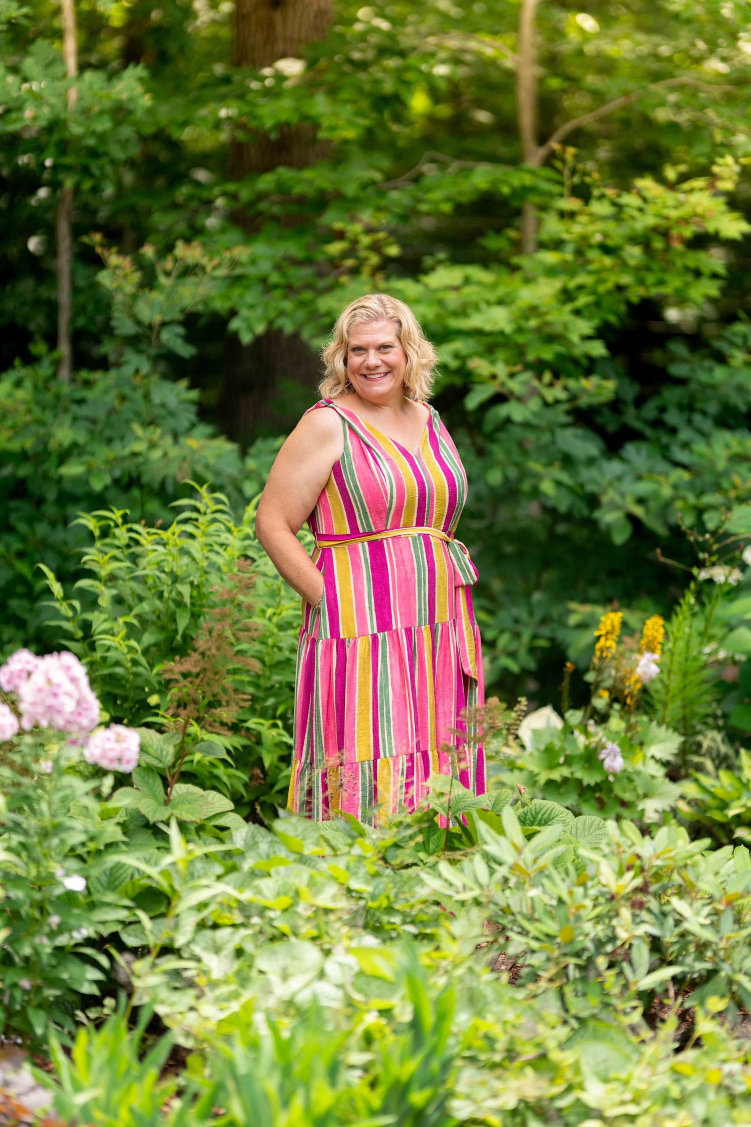 A smiling blonde woman in a colorful striped dress stands among lush green garden foliage.