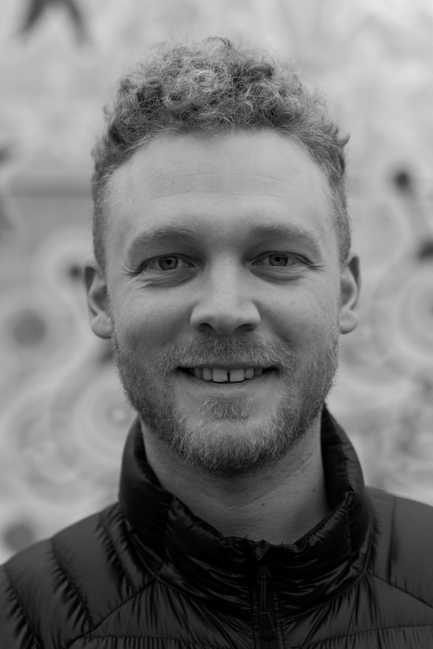 Black and white portrait of a smiling young man with curly hair and a beard outdoors.