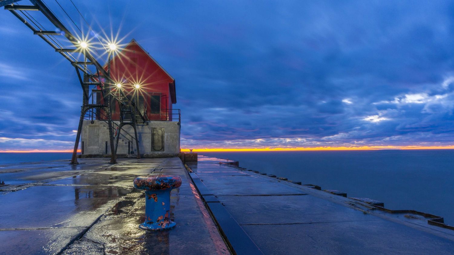 Red lighthouse building on a wet pier glowing under blue cloudy sky at dusk over calm water.