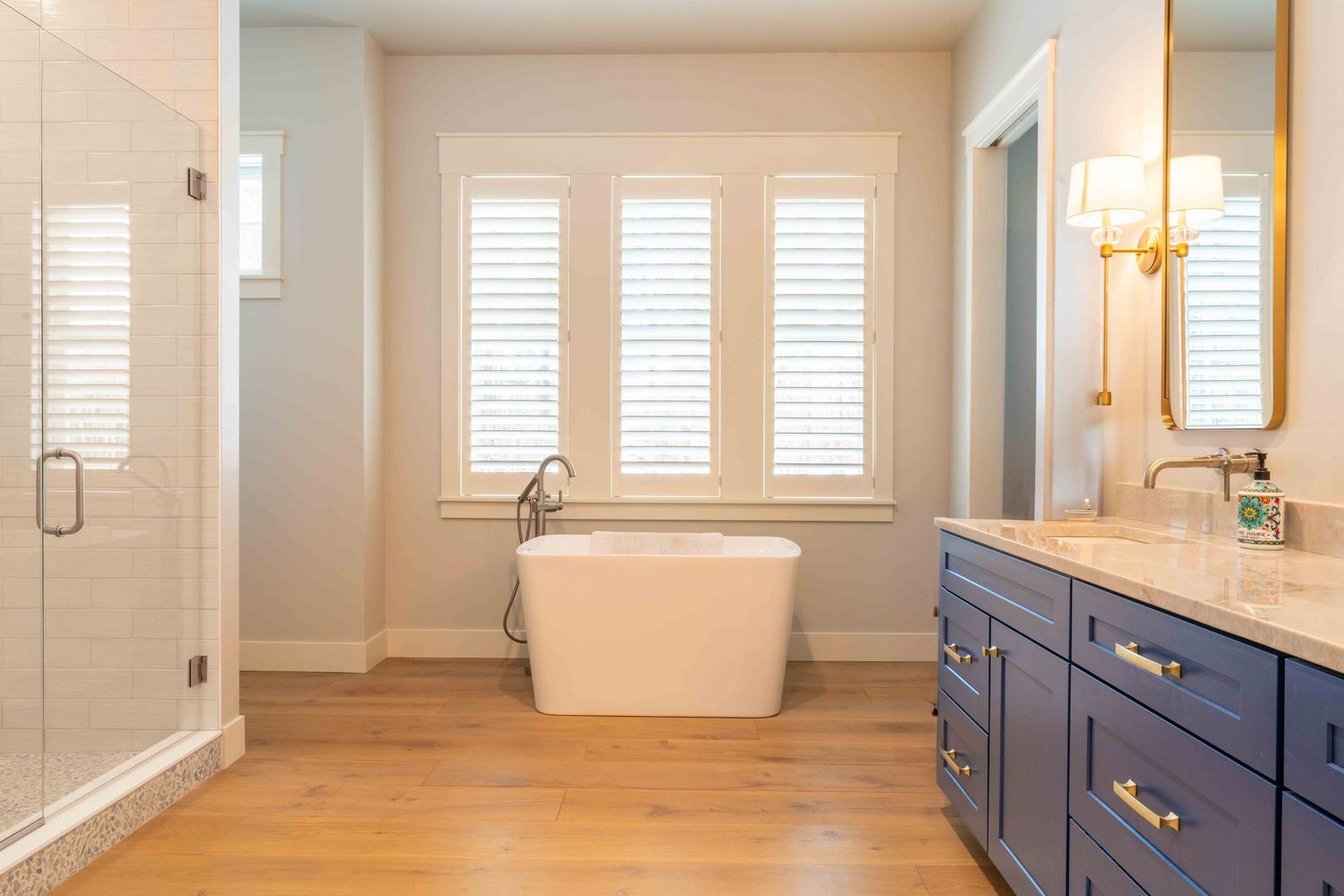 Spa-like bathroom with navy vanity, freestanding soaking tub, glass shower, and plantation shutters.