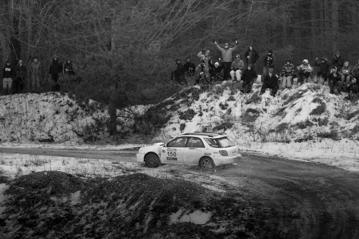 Rally car navigates a snowy dirt road curve with spectators watching from hillside.