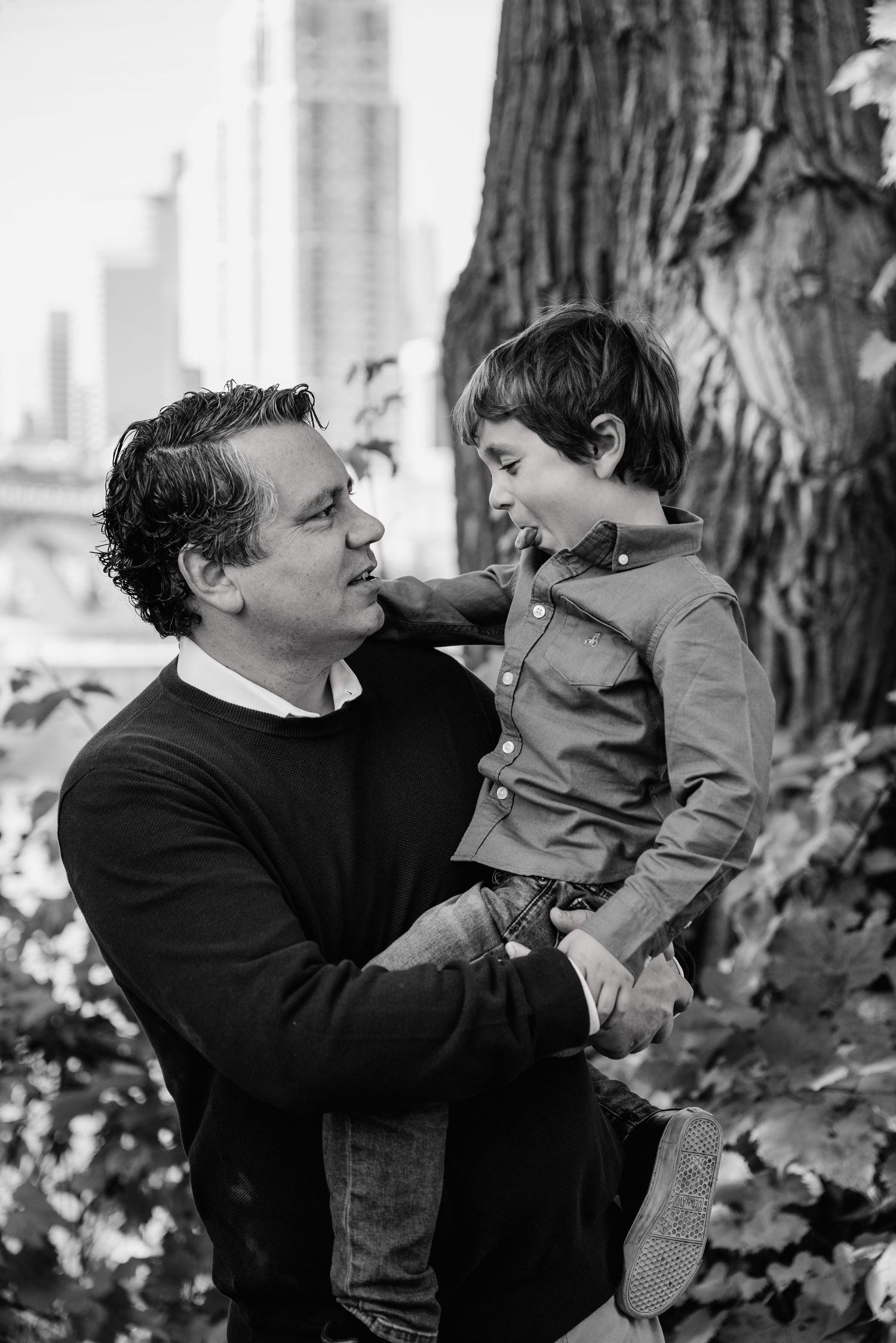 Black and white portrait of a father holding his young son beside a large tree with city buildings behind.