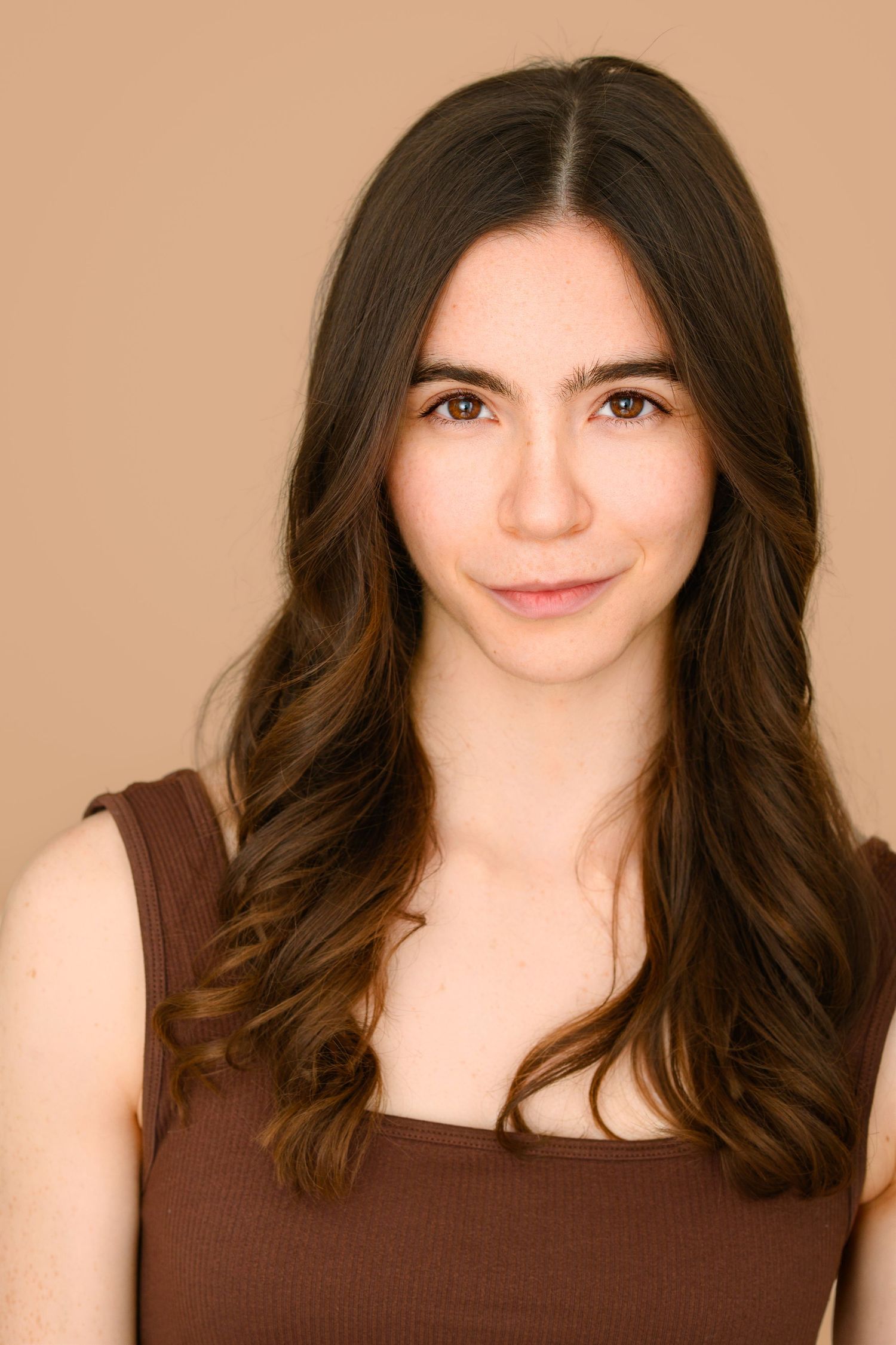 Headshot of a young woman with long wavy brown hair and brown eyes wearing a brown sleeveless top against a tan background.