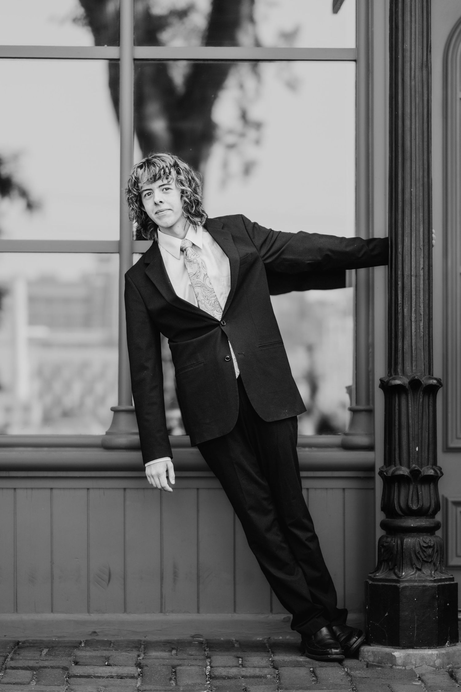 Black and white portrait of a curly-haired young man in a dark suit and tie leaning against a column outside a window.