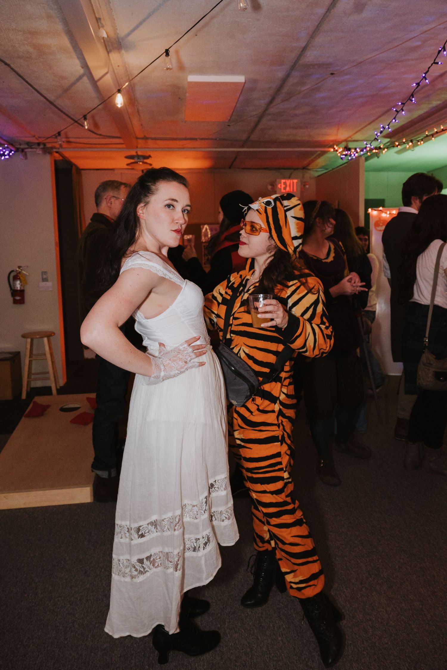 Two women pose at a costume party, one in a white lace dress and the other in a tiger-striped jumpsuit, holding a drink under string lights.