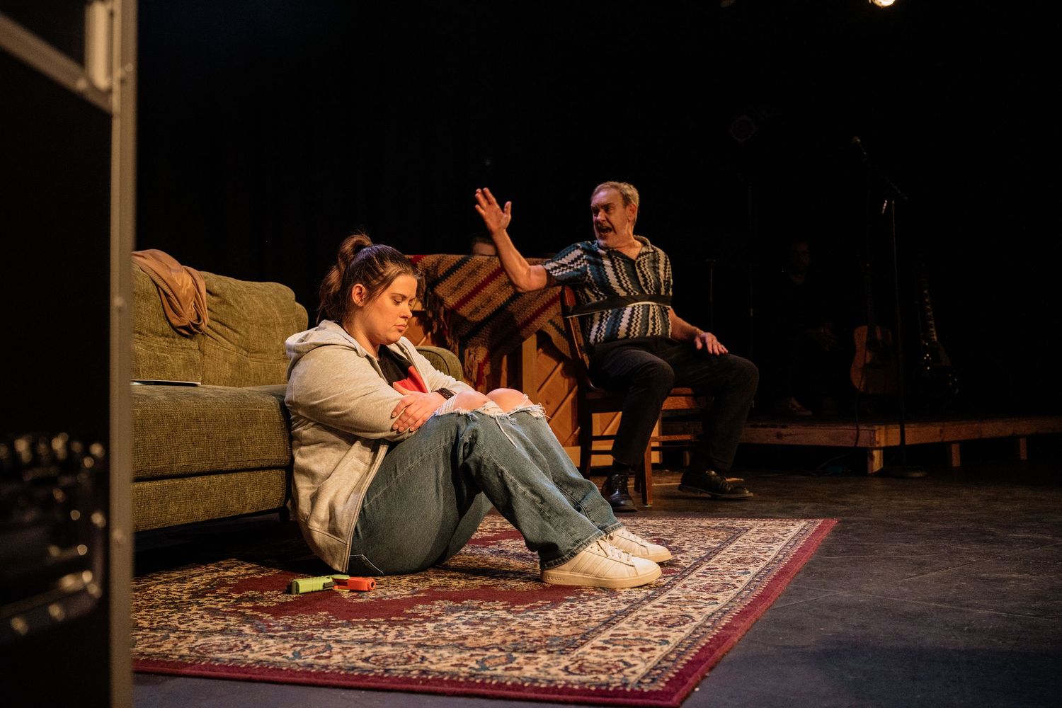 A woman sits on a rug beside a green couch while a man gestures from a chair during a theater rehearsal on stage.