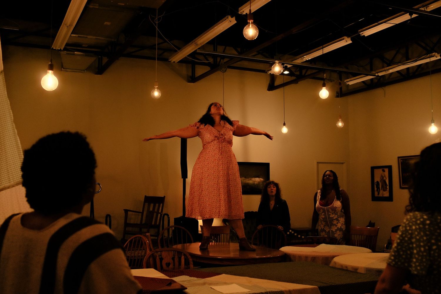 A performer in a pink dress stands on a raised platform with arms spread wide, lit by warm hanging bulbs as an audience watches.