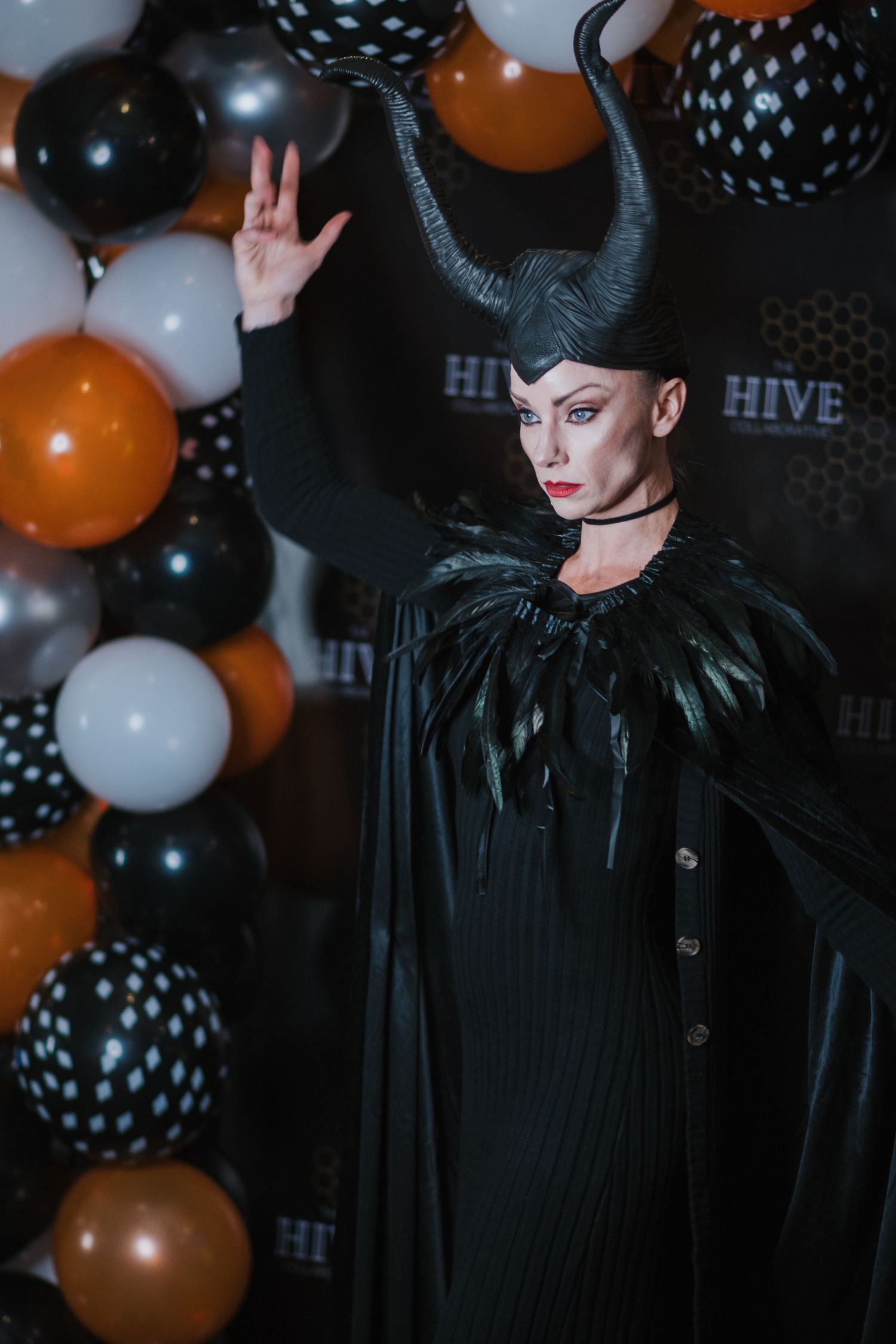 Woman in a Maleficent-style Halloween costume with black horns and feathered cape poses beside orange, black and white balloons.