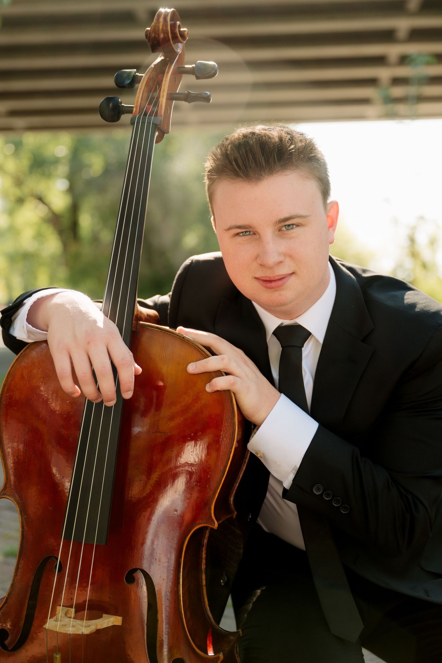 Young musician in a black suit and tie leans on his cello while posing outdoors under a covered walkway.