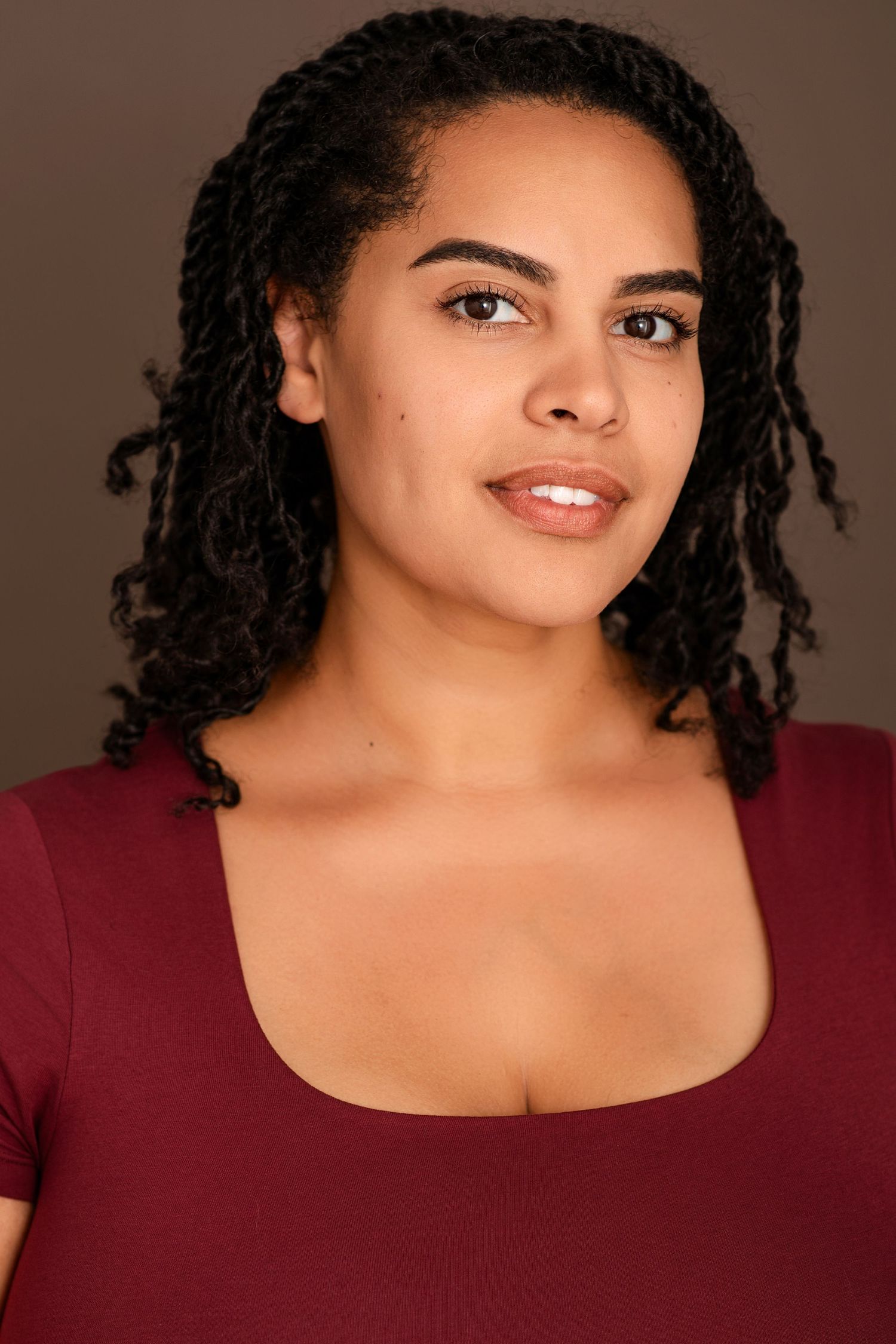 Headshot of a smiling woman with twisted black hair wearing a burgundy scoop-neck top against a brown background.