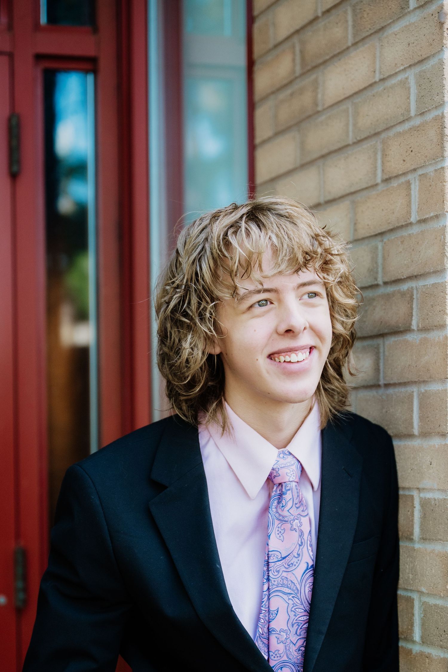 Smiling young man with wavy shoulder-length hair wears a dark blazer and paisley tie beside a red door and brick wall.