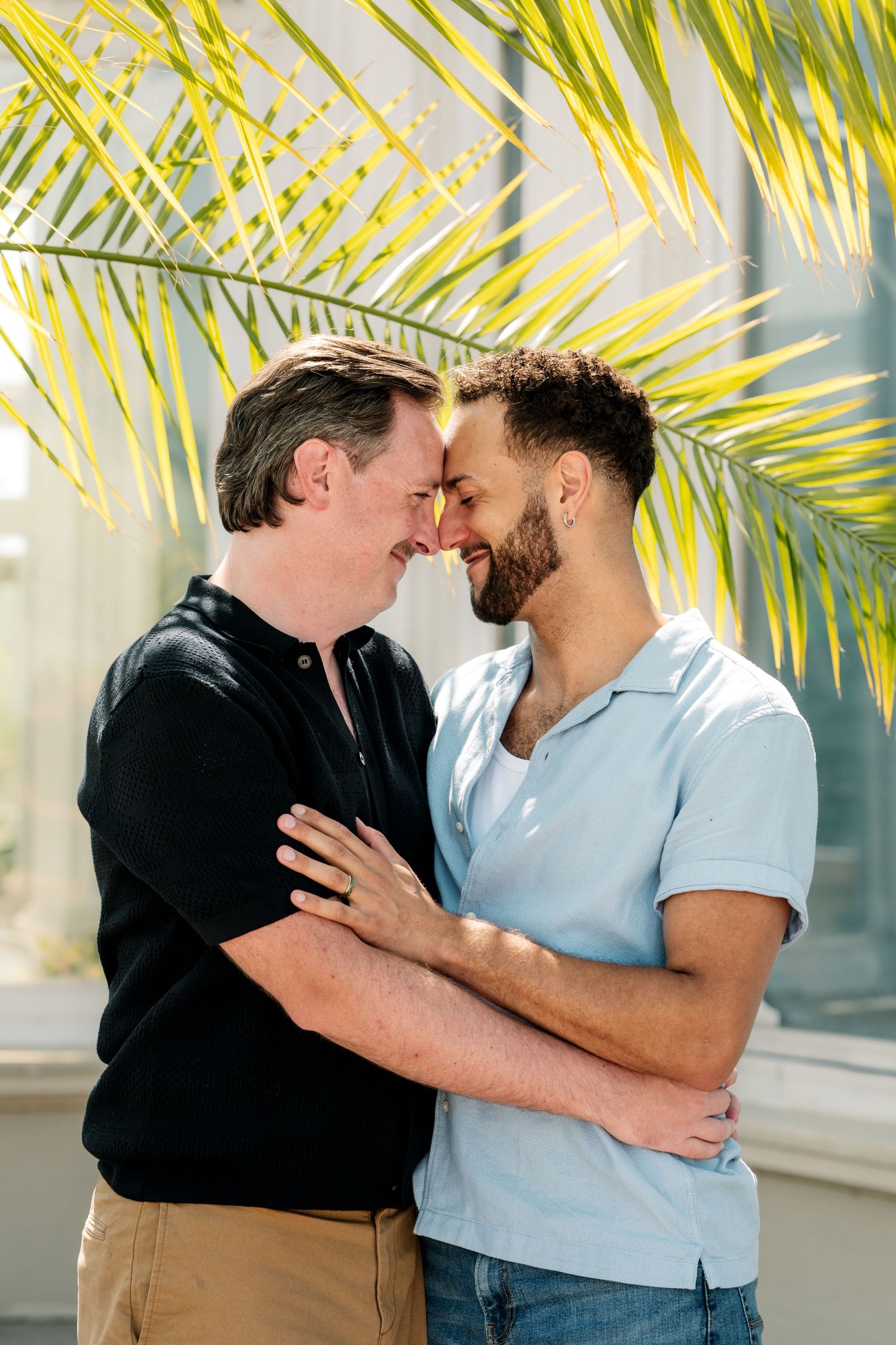 Two men touch foreheads and smile in a warm embrace beneath yellow-green palm fronds on a bright sunny day.