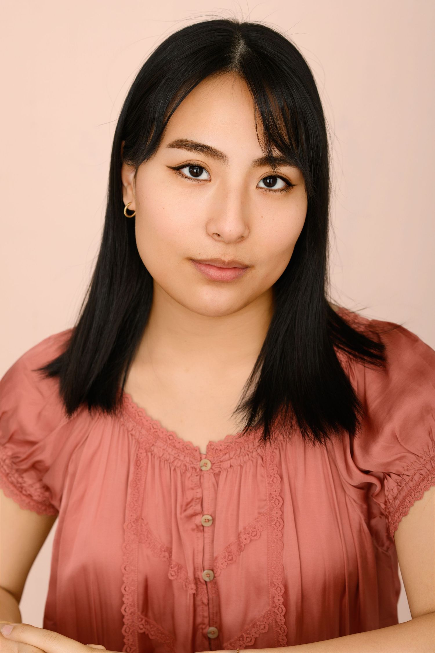 Headshot of a young woman with straight black hair and bangs wearing a coral lace-trimmed blouse against a pink background.