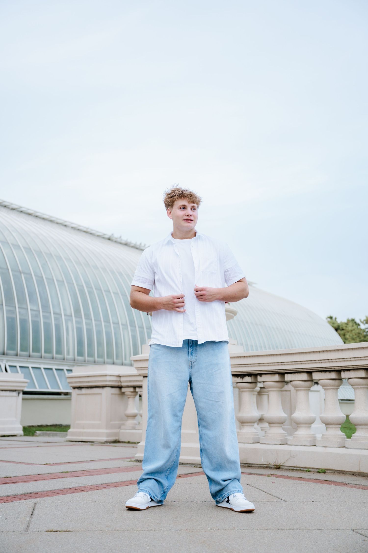 Young man in a white shirt and light blue jeans stands on a stone terrace in front of a large glass conservatory.
