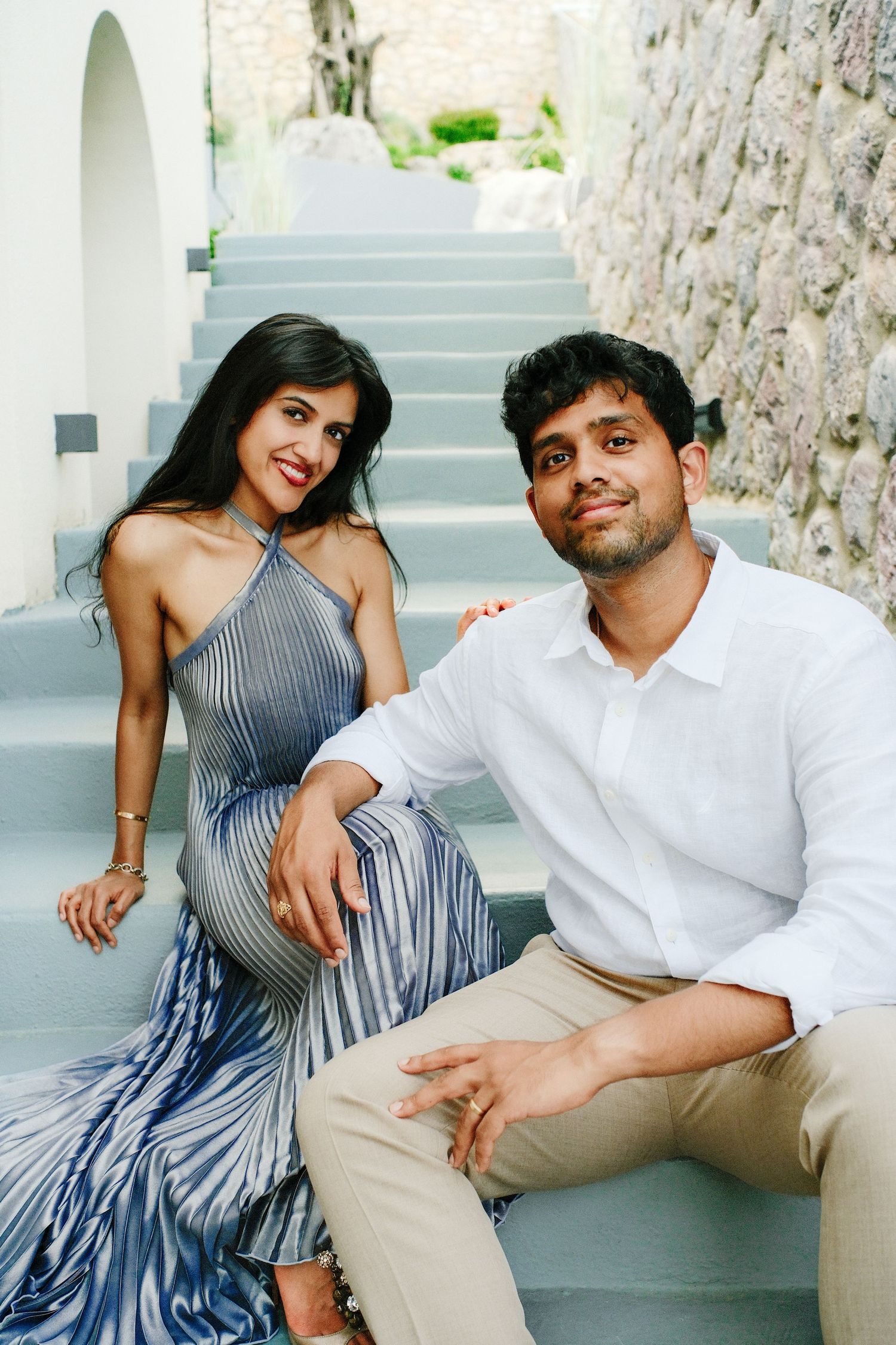 A smiling couple sits together on pale stone steps, she in a pleated blue gown and he in a white shirt and khakis.