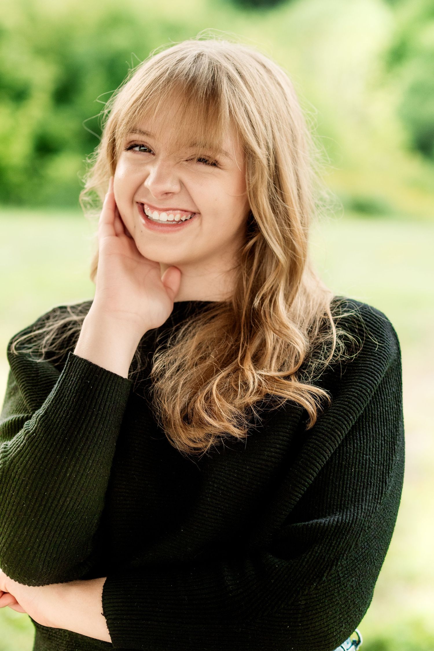 Smiling young woman with long wavy blonde hair and bangs wears a dark green sweater and rests her cheek on her hand outdoors.