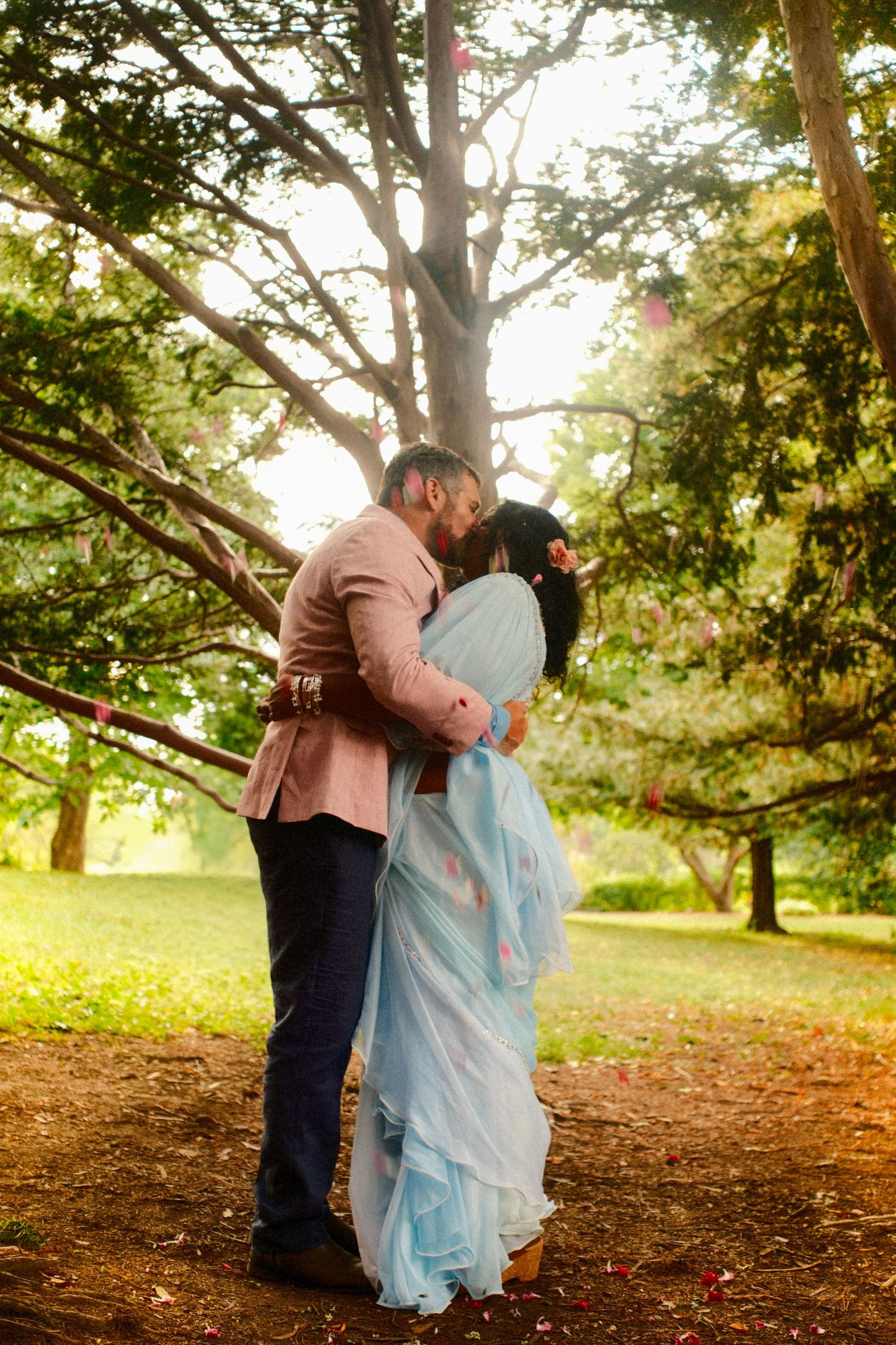 A couple in a pink blazer and light blue sari share a kiss beneath tall trees in a sunlit park with scattered petals on the ground.