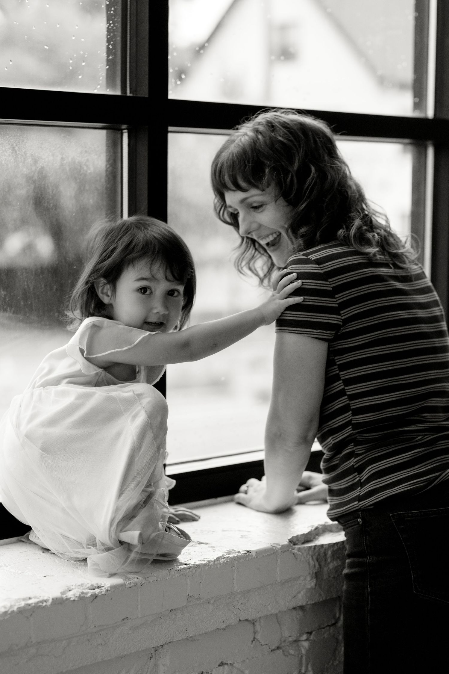 Black and white photo of a smiling mother and her toddler daughter playing by a rain-spotted window.