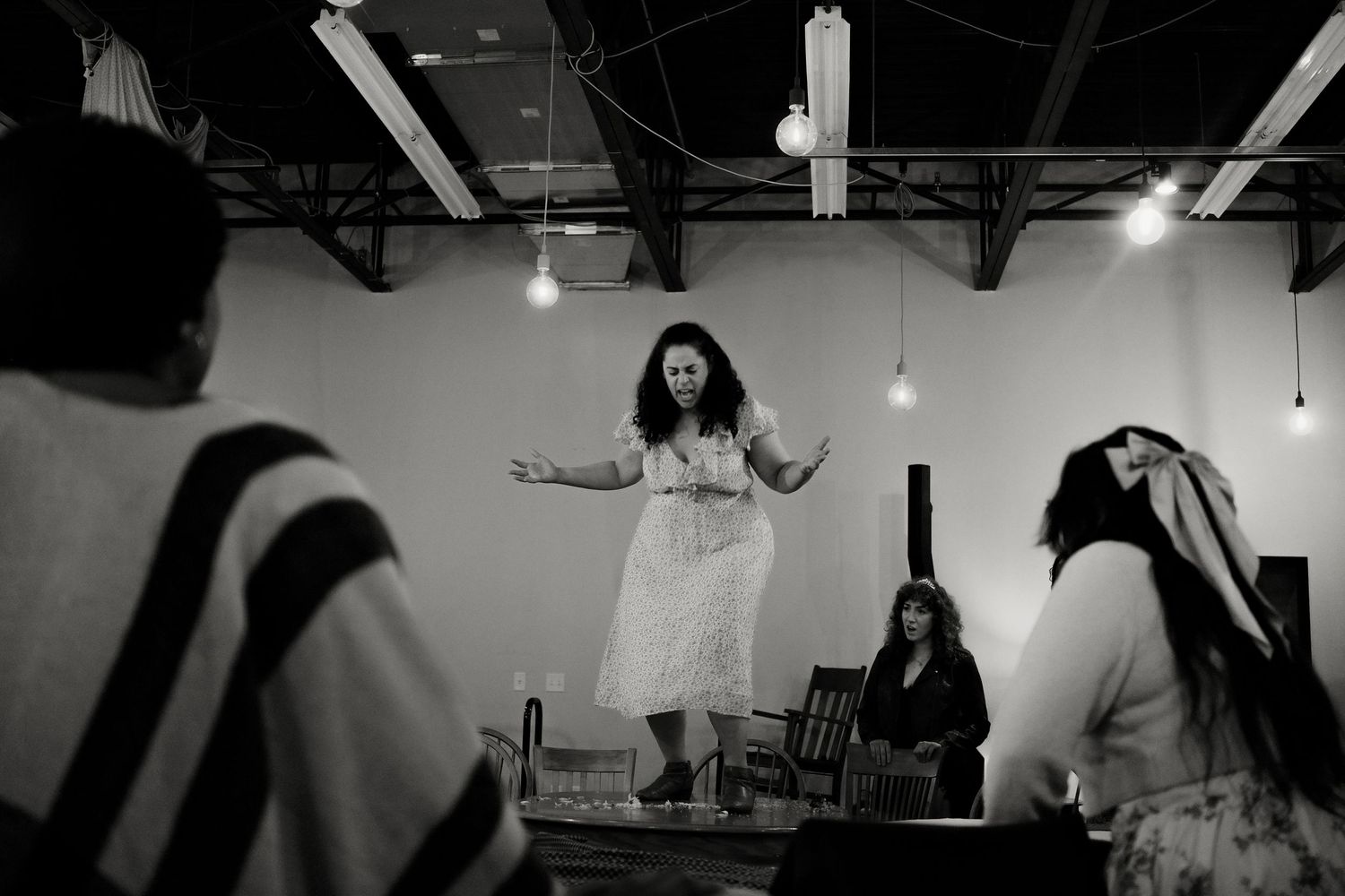 A woman in a white dress stands with arms outstretched on a small stage as audience members watch in a rustic, industrial space.