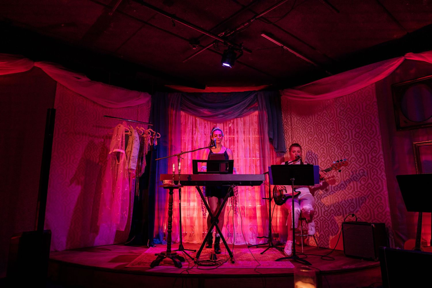 Two musicians perform on a small stage with a keyboard and guitar, framed by glowing pink and blue draped curtains in an intimate venue.