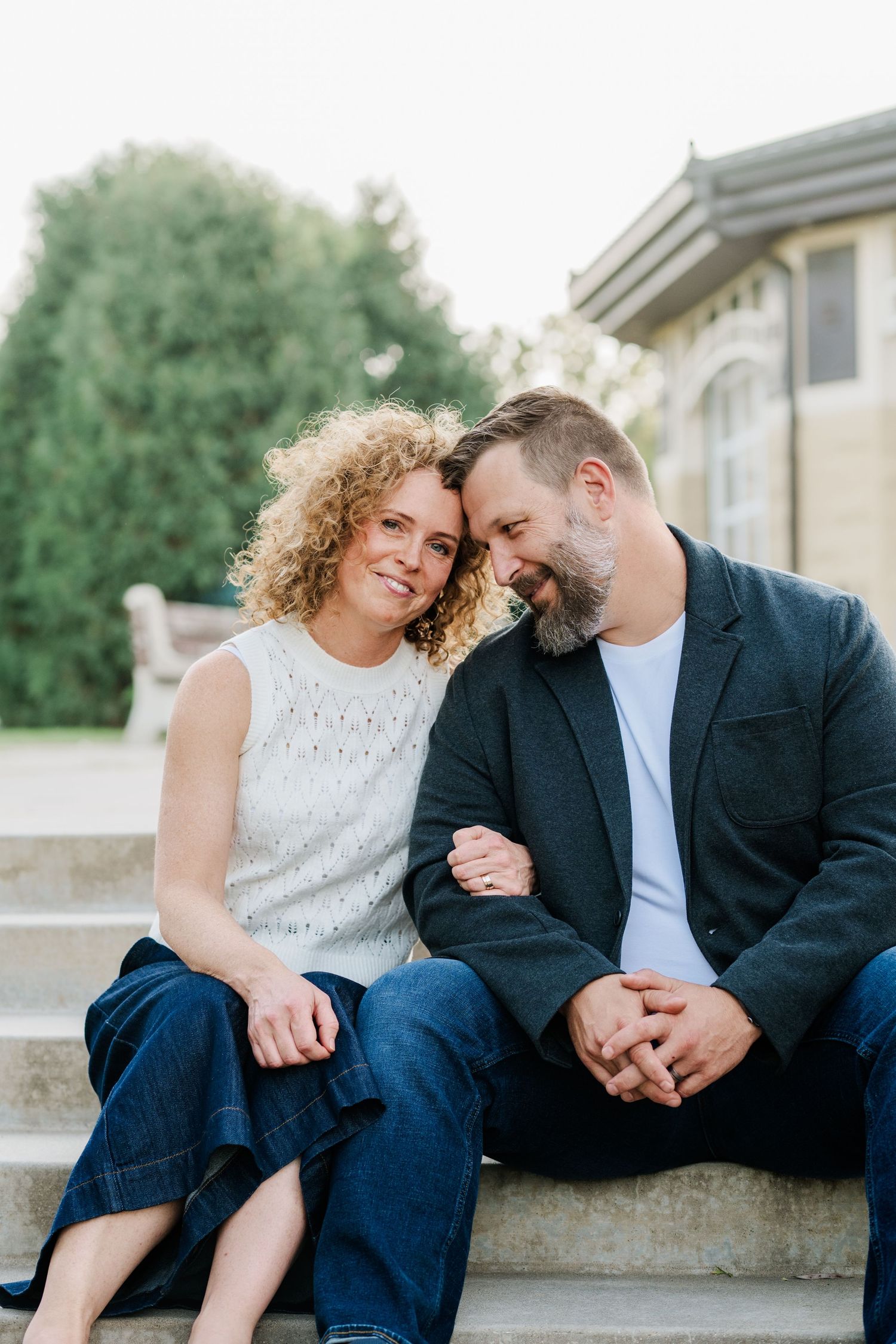 A smiling curly-haired woman and a bearded man lean together on stone steps in a warm outdoor couple portrait.