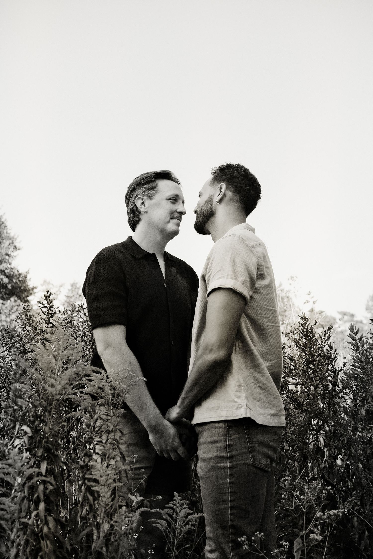 A black and white portrait of two men holding hands and gazing at each other among tall wild plants outdoors.