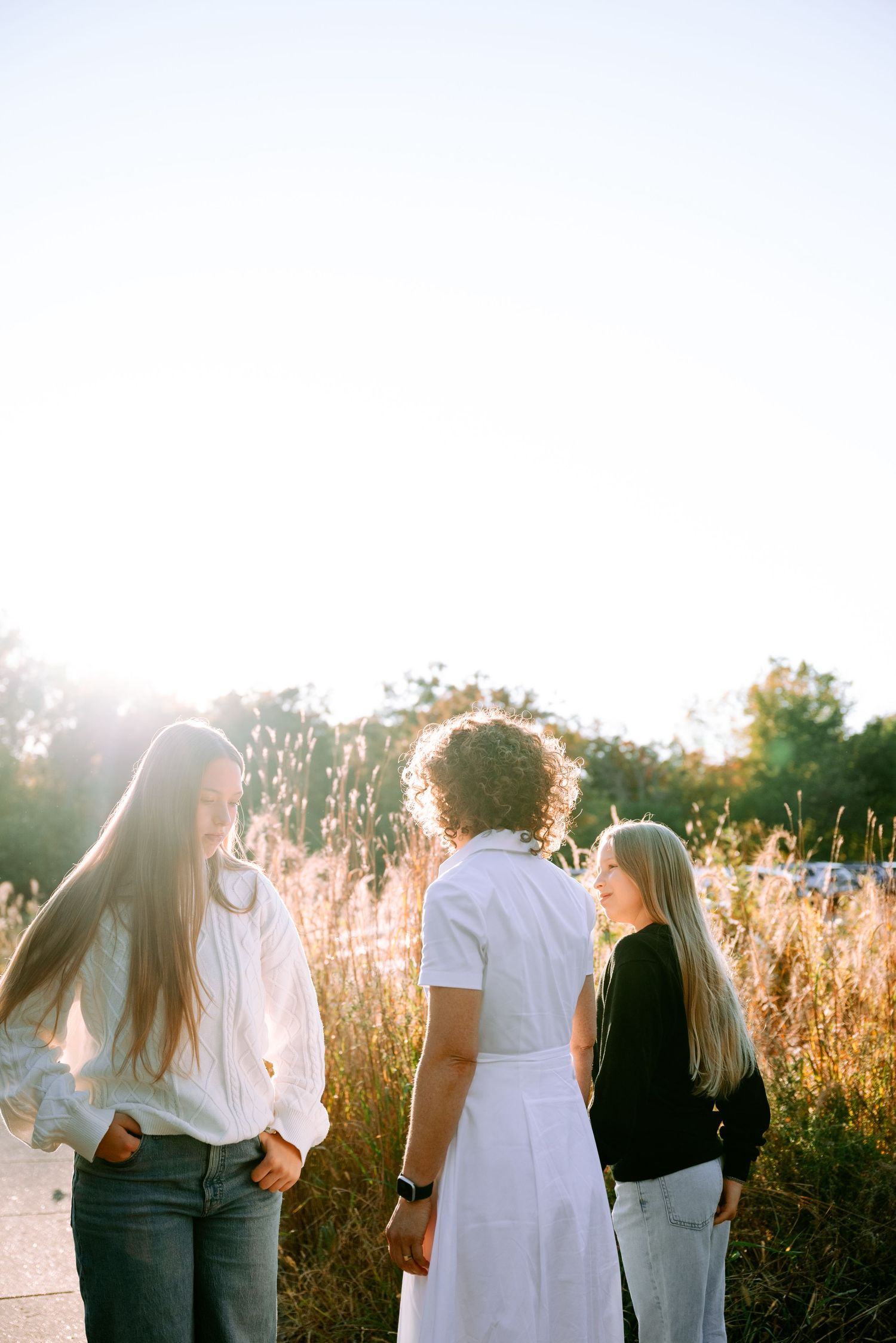 Three girls stand in golden sunlight among tall grasses in a bright, airy outdoor portrait.