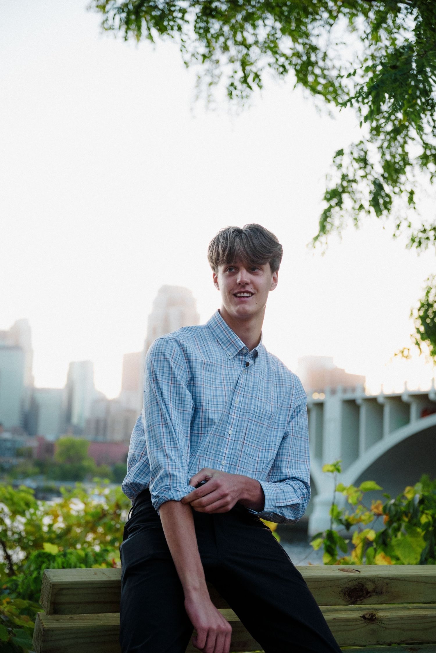 Young man in a blue plaid shirt sits on a wooden bench with a city skyline and bridge in the background.