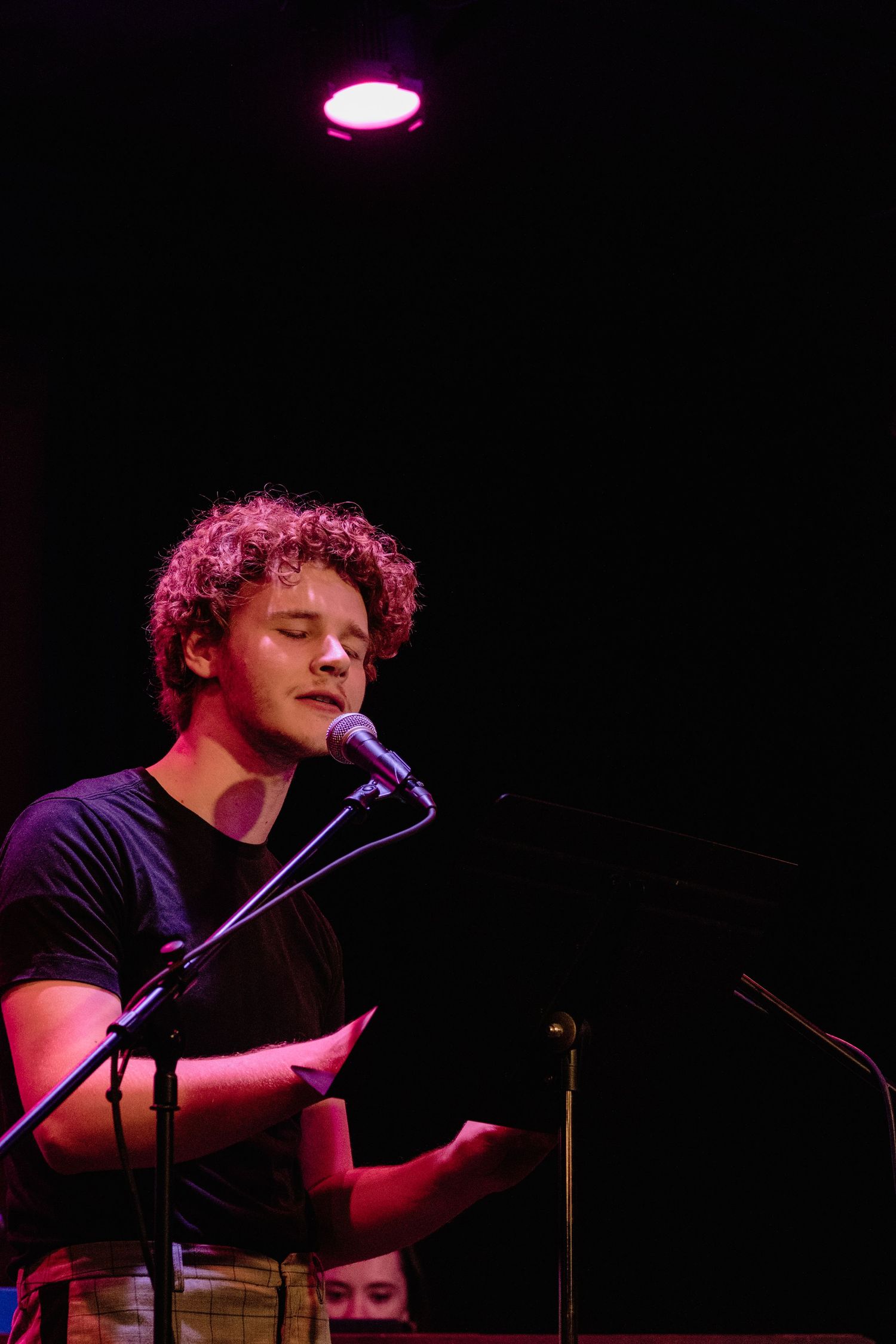 A curly haired young man in a dark t-shirt speaks into a microphone under a pink stage light, holding papers.