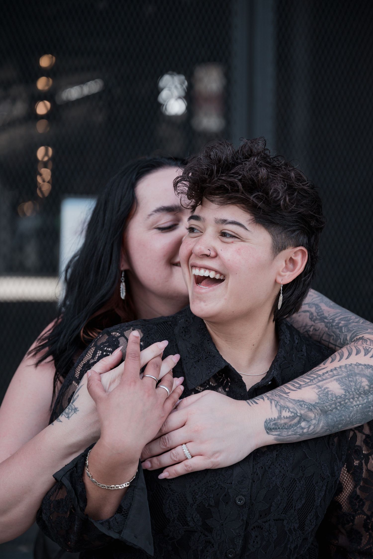 A tattooed person laughs joyfully while a dark-haired partner kisses their cheek in a playful, affectionate couple portrait.
