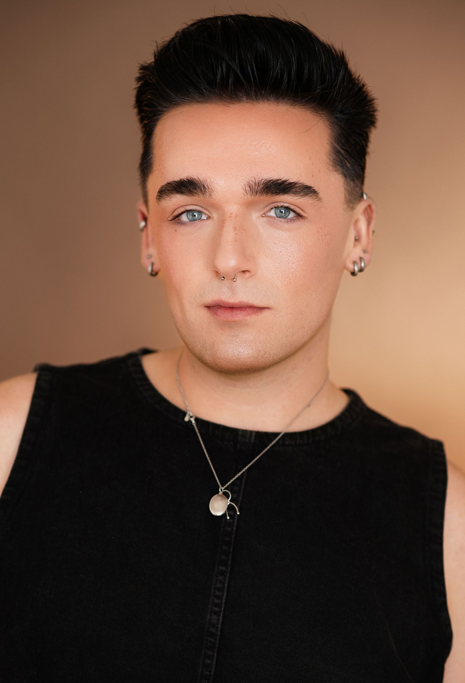Young person with styled black hair, ear piercings, and a pendant necklace wears a black sleeveless top against a warm brown background.