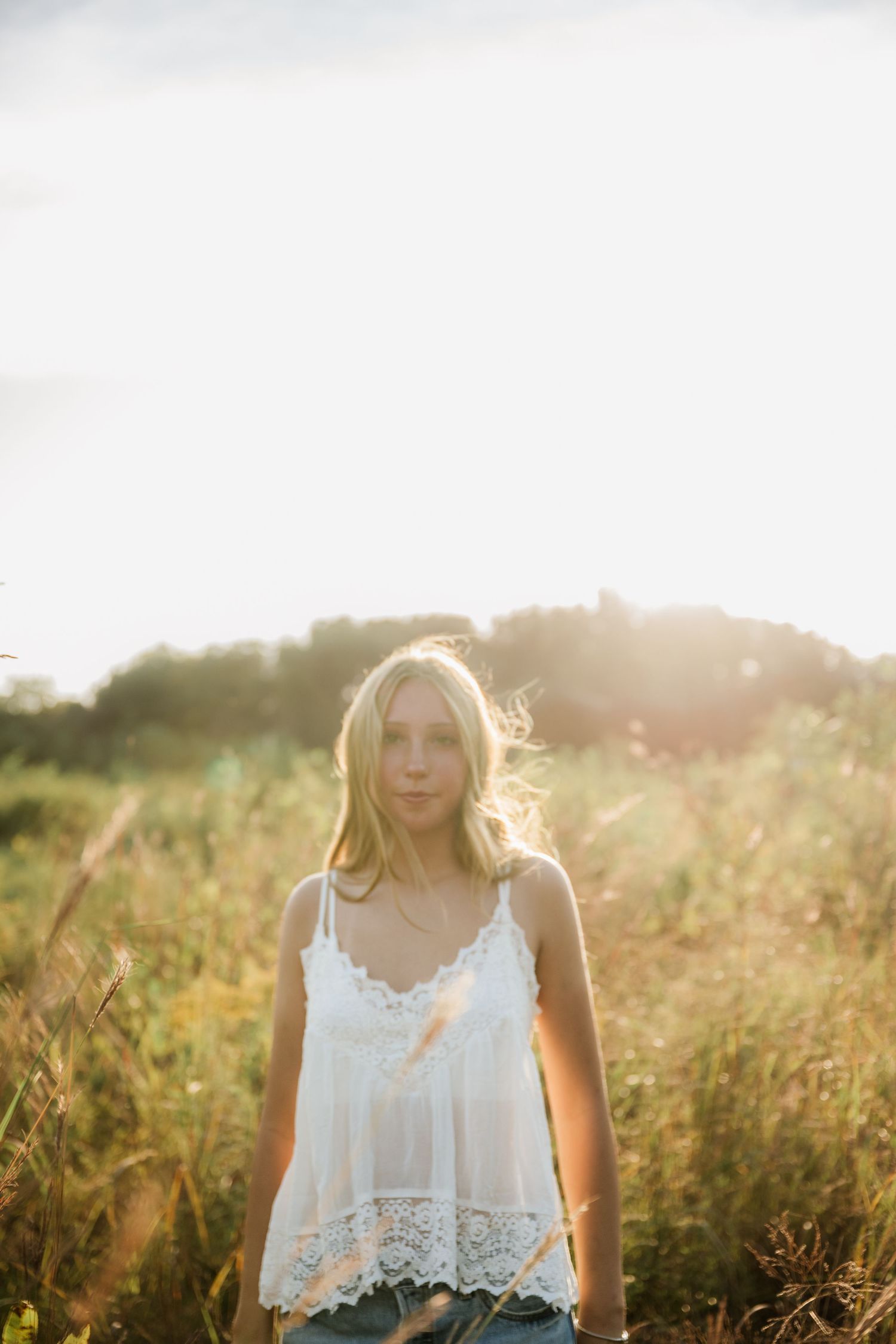 Young woman with long blonde hair stands in a sunlit field of tall golden grass wearing a white lace camisole.