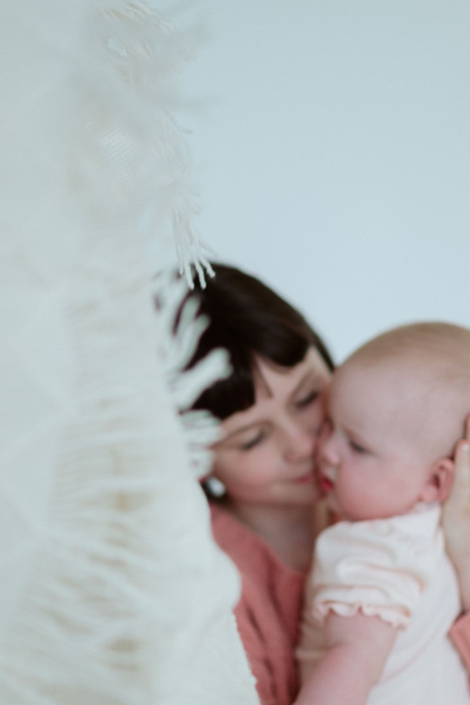 A dark-haired child in a coral top cuddles her baby sister in soft focus, with a white feathered object blurred in the foreground.