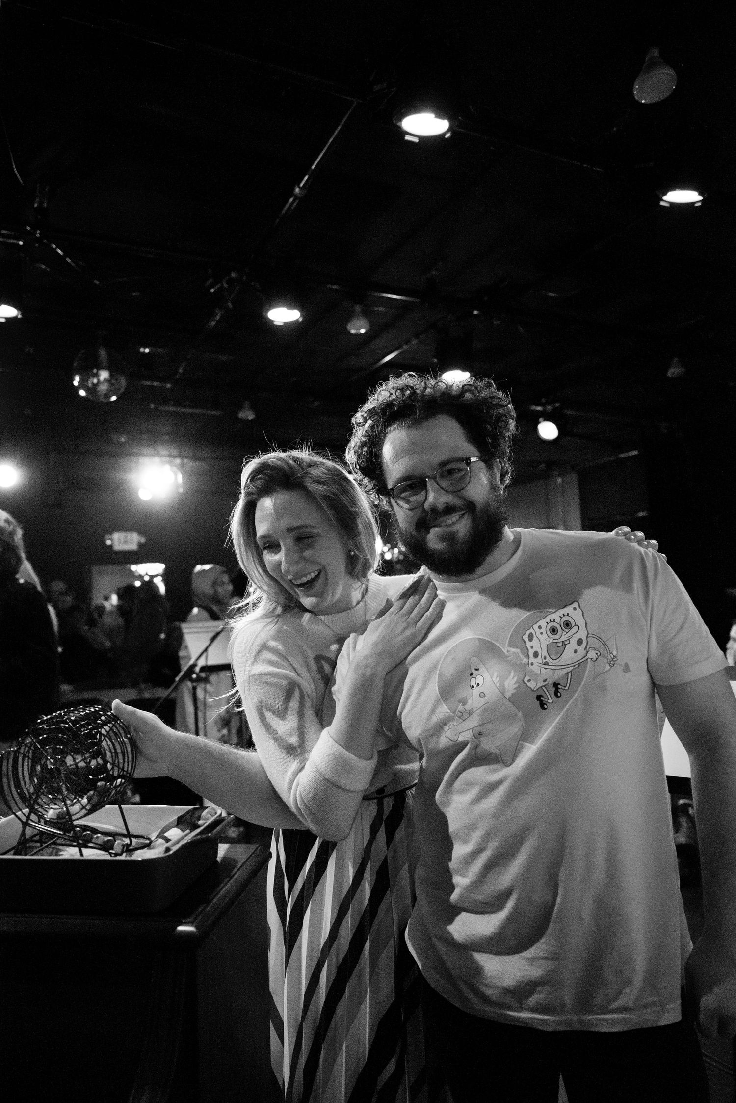 A smiling woman leans against a bearded man in glasses wearing a graphic tee, standing beside a bingo cage in a dimly lit venue.