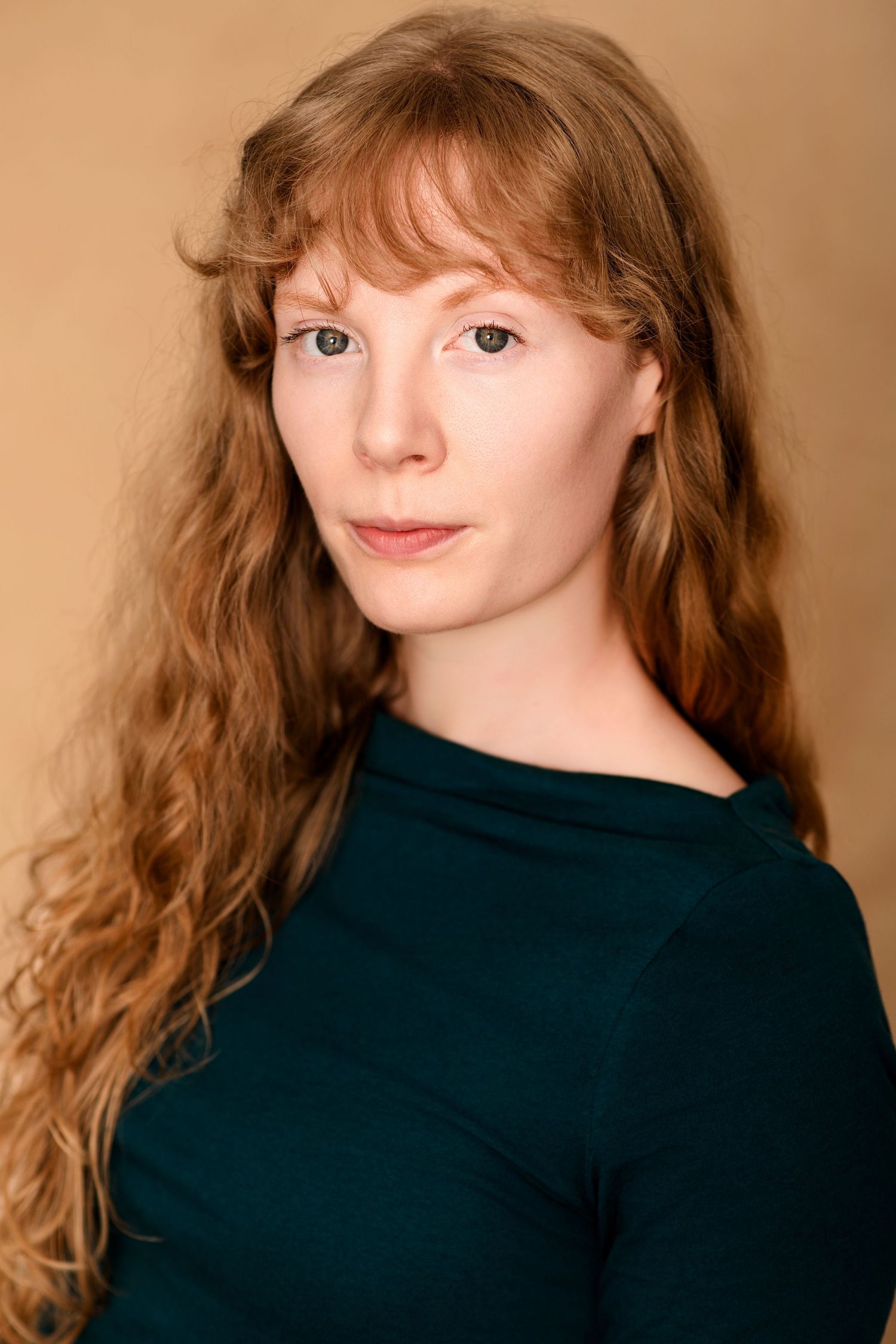 Headshot of a woman with long wavy auburn hair and bangs wearing a dark green mock-neck top against a tan background.