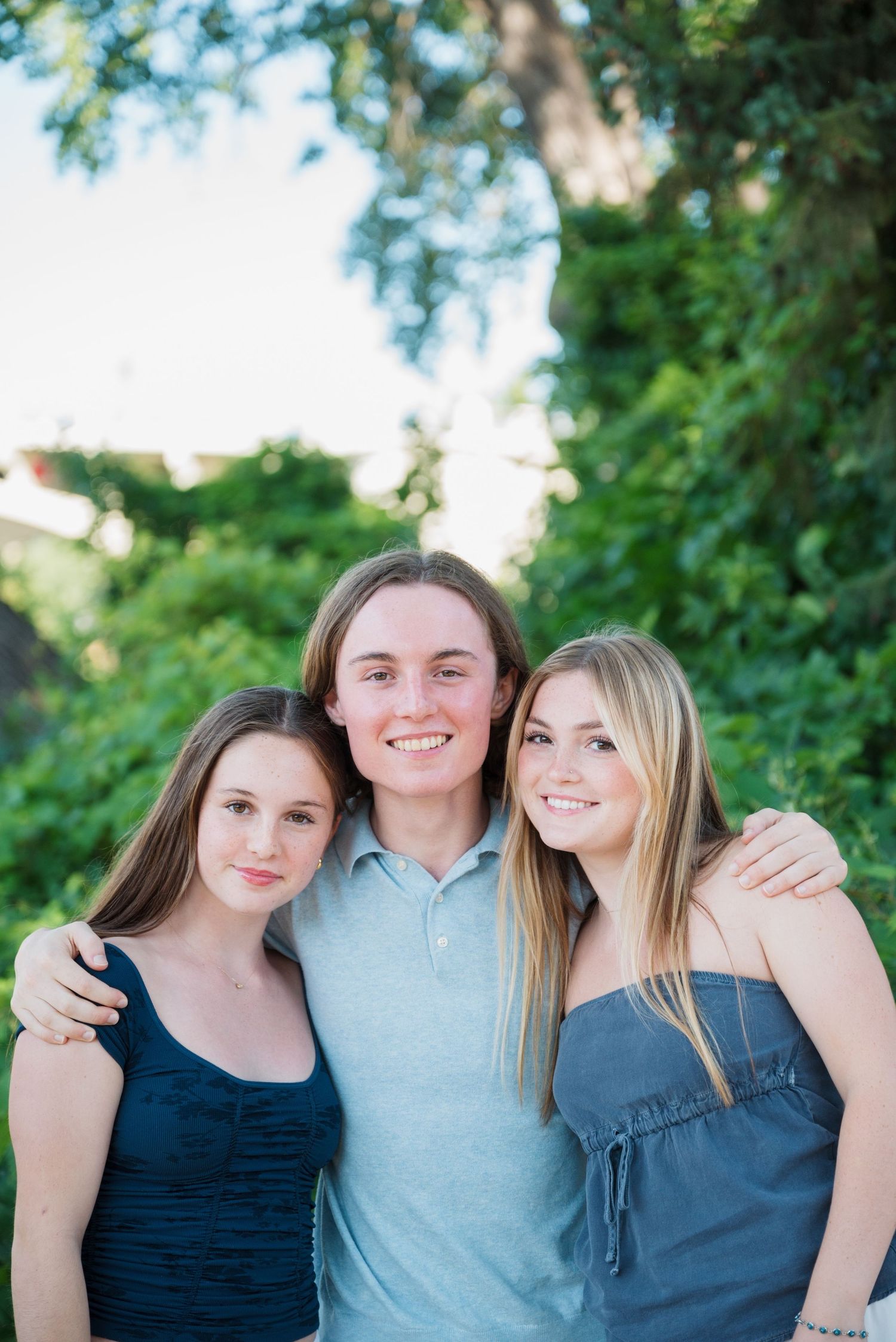 Three smiling young people hug and pose together outdoors in front of lush green foliage during a sunny family photo session.