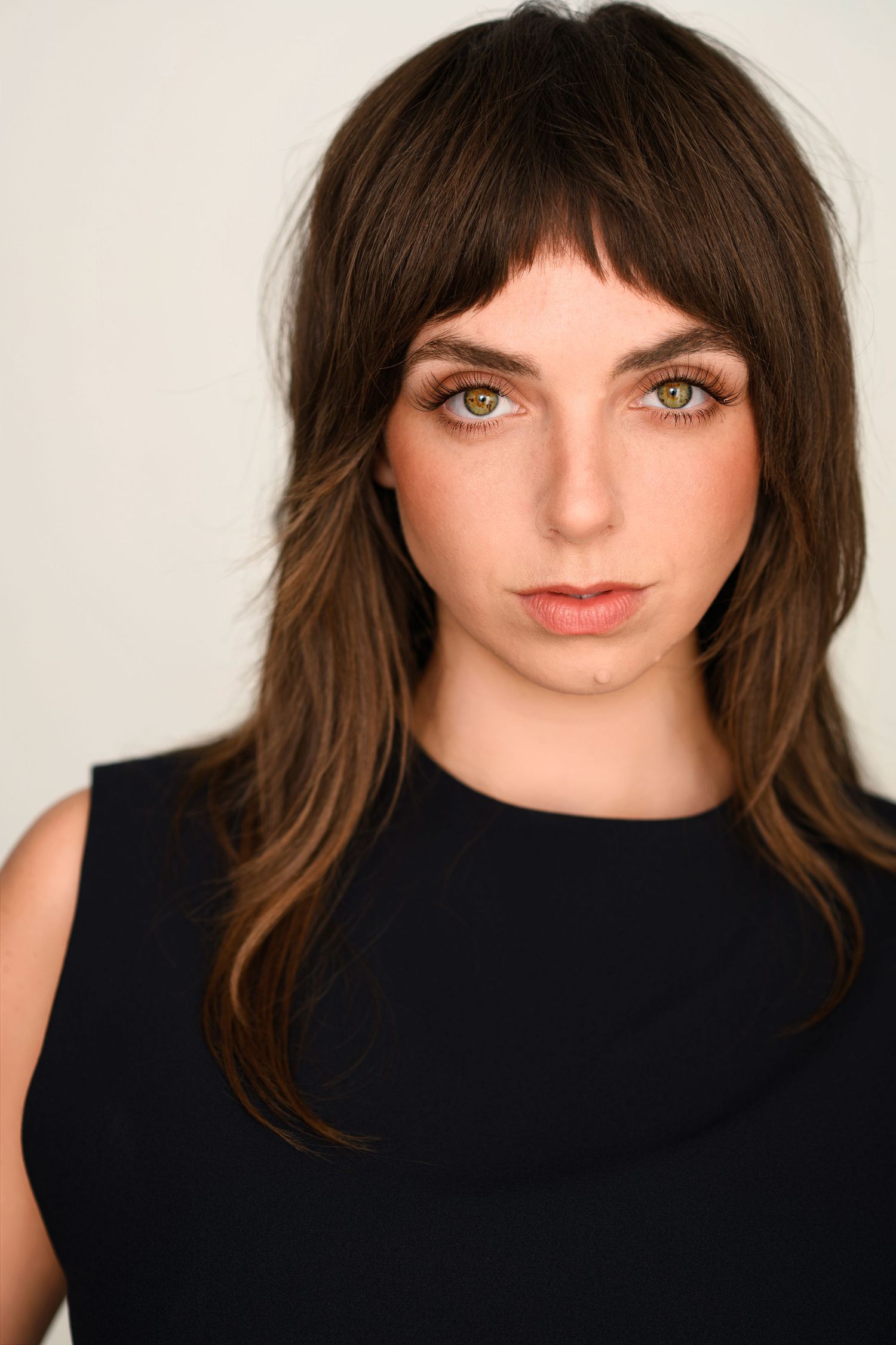 Headshot of a young woman with brown shaggy layered hair, bangs and hazel eyes wearing a black sleeveless top.
