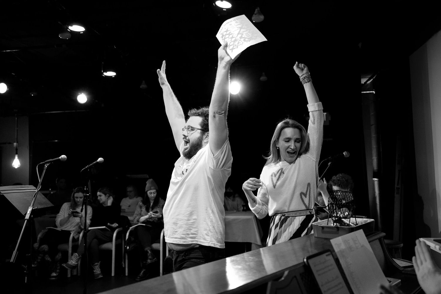 A man and a woman cheer with arms raised in triumph on a stage while holding papers, in black and white.