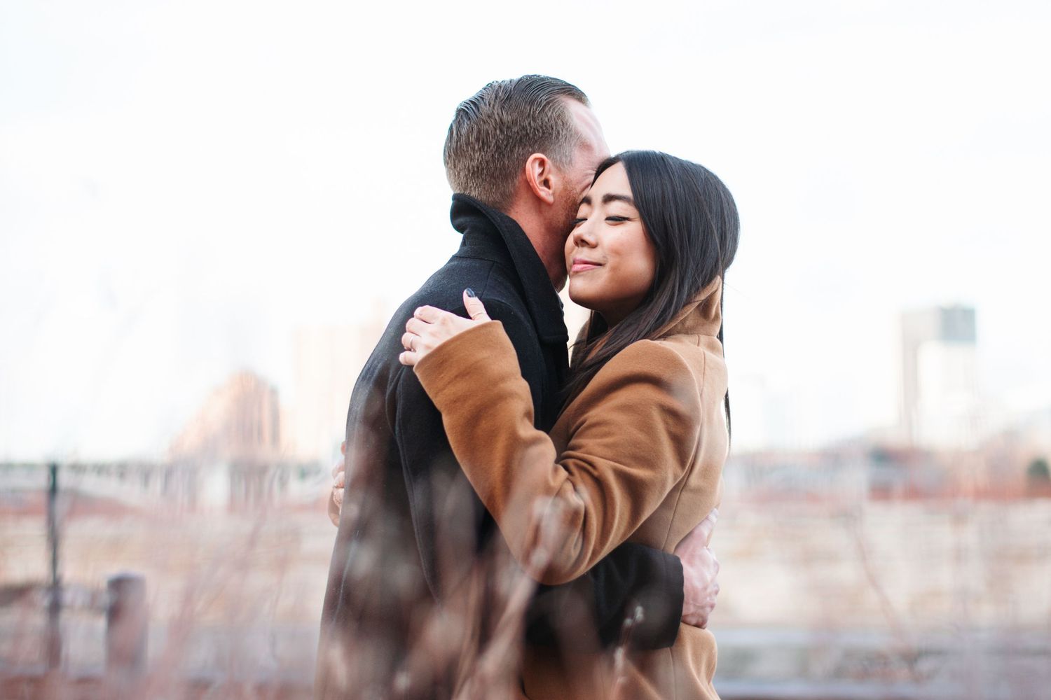 A couple in winter coats shares a tender embrace on a city rooftop, the woman resting her head against the man's cheek.